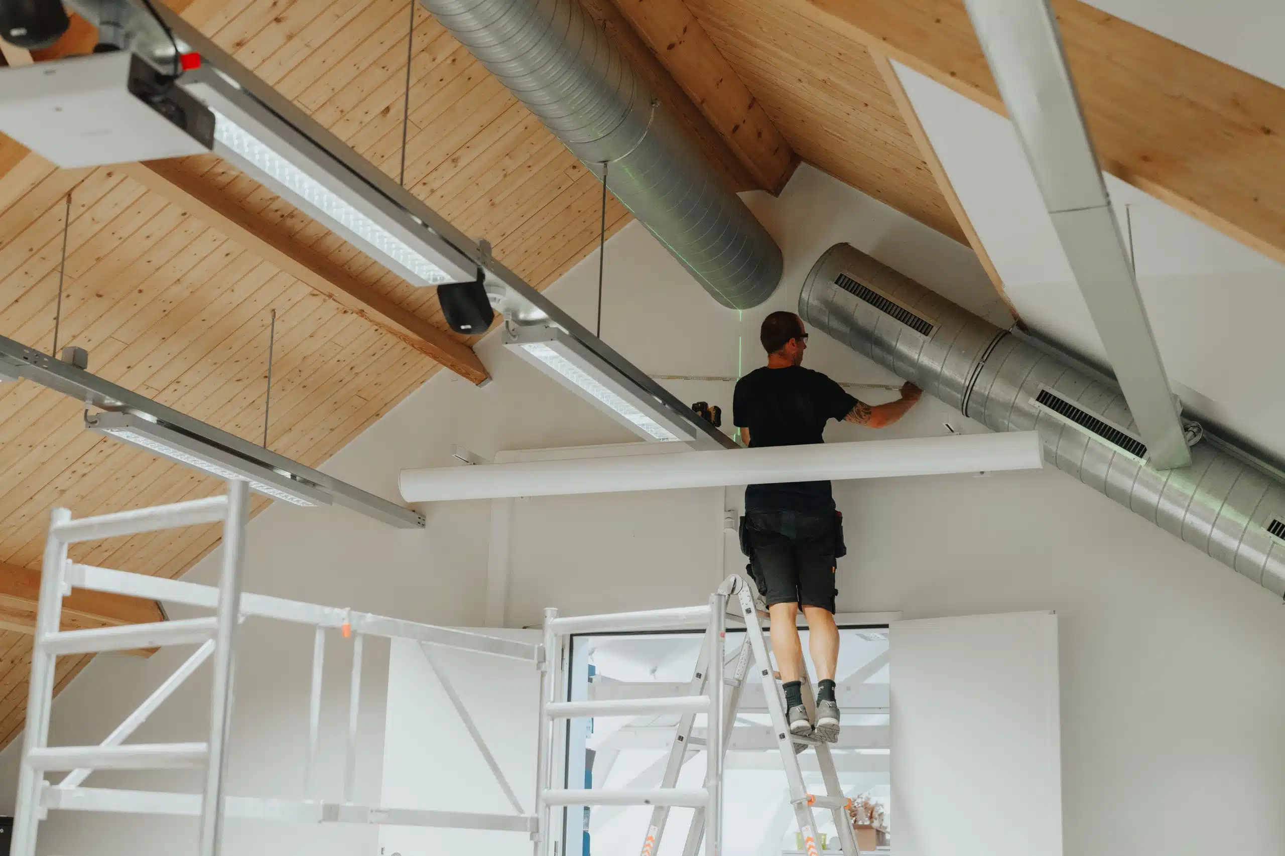 Installateur monteert ventilatie-unit onder plafond met houten balken en leidingen